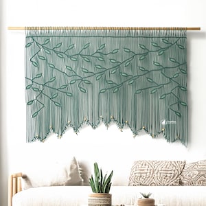 Puede incluir: Un tapiz de macramé verde con un diseño de hojas. La obra de arte está suspendida de una barra de madera y tiene un borde inferior festoneado con pequeñas cuentas de color claro. Esta pieza decorativa añade un toque natural y bohemio a cualquier habitación.