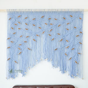 Pode incluir: Uma tapeçaria de macramé azul claro com um design de folhas. As folhas são feitas de cordão marrom e a tapeçaria é adornada com contas de madeira.