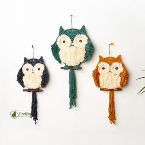 May include: Three owl-shaped wall hangings in navy, green, and orange, each with cream-colored faces and dangling tassels. The owls are displayed above a beige sofa with decorative pillows, a woven rug, and wooden tables.