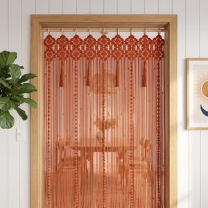 May include: A burnt orange macrame door curtain featuring an intricate knotted top panel with repeating geometric patterns and long, vertical fringe strands. The curtain is suspended from a wooden rod mounted within a wooden door frame.