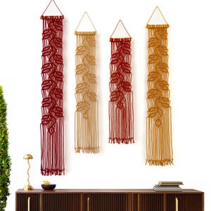 May include: Four vertical macrame wall hangings suspended from wooden dowels with triangular hanging cords. Two hangings are deep red and two are mustard yellow, each featuring a repeating knotted pattern that ends in long, loose fringe.