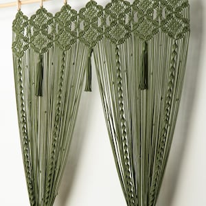 May include: Olive green macrame curtain panel with a geometric top design and tassels. The curtain hangs from a wooden rod with wooden rings. The curtain is made of many vertical strands of knotted cord.