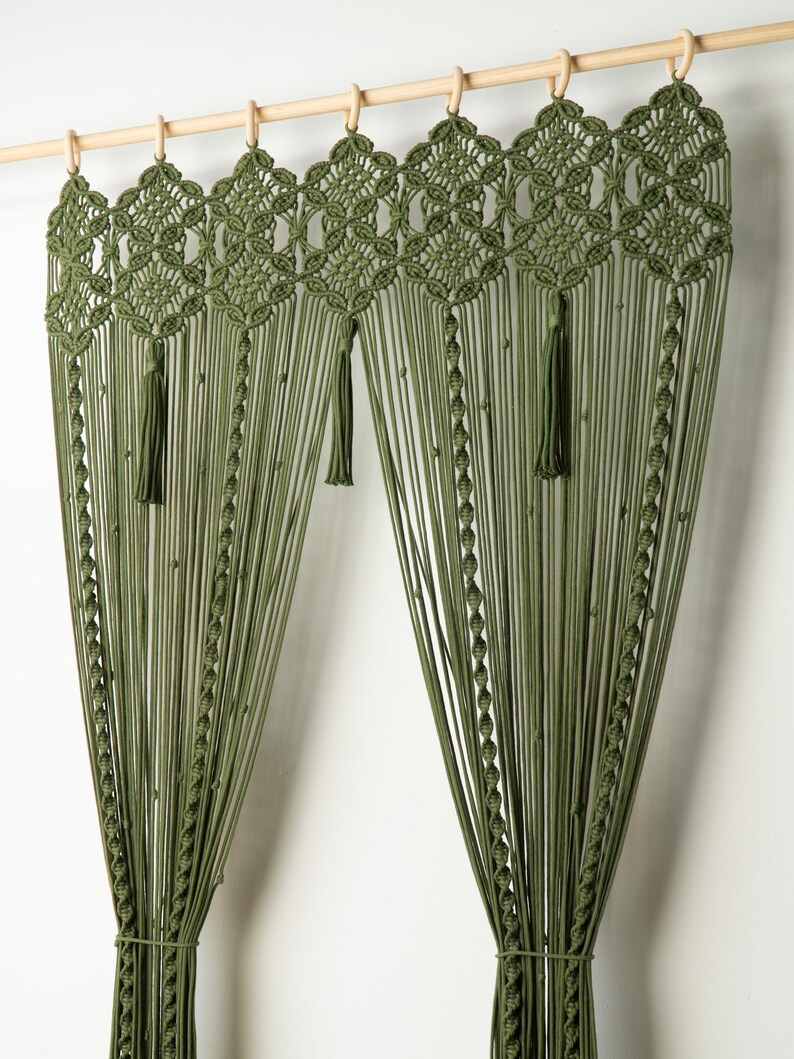 May include: Olive green macrame curtain panel with a geometric top design and tassels. The curtain hangs from a wooden rod with wooden rings. The curtain is made of many vertical strands of knotted cord.