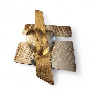 May include: Abstract metal brooch with a gold vertical bar intersecting a silver heart and textured silver rectangles. The brooch has a modern, geometric design with a mix of polished and textured surfaces. The brooch is a unique piece of jewelry.