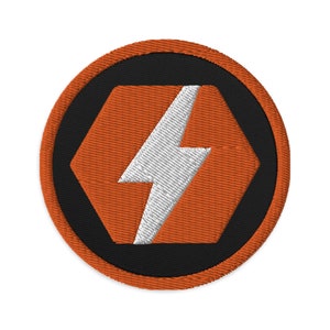 以下が含まれることがあります： オレンジと黒の刺繍パッチで、六角形の中に白い稲妻が描かれています。