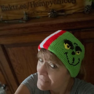 May include: A green crocheted hat with a red and white stripe and a Grinch face design. The hat is being worn by a person with short brown hair.