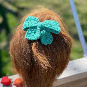 May include: A teal crocheted bow hair tie. The bow is tied in a knot and has a small pom-pom in the center.