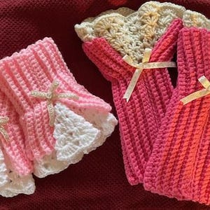 May include: Handmade crochet baby booties and bottle covers in pink and cream. The booties have a white lace trim and are adorned with a small bow. The bottle covers are a vibrant pink with a cream bow.