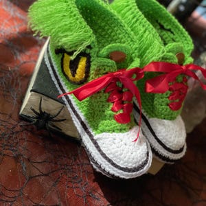 Puede incluir: Zapatillas de ganchillo verdes hechas a mano con suelas blancas y ribetes negros. Las zapatillas presentan un diseño de personaje de dibujos animados con un ojo amarillo y cordones de cinta roja. Un artículo de calzado único y caprichoso.