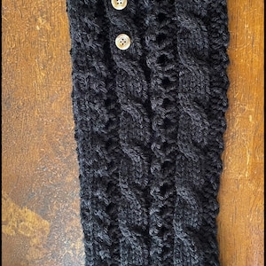 Puede incluir: Calentadores de piernas de punto negro con un patrón de punto de cable y tres botones blancos. Los calentadores de piernas tienen una etiqueta de cuero marrón con el texto "The Little Knit Co."