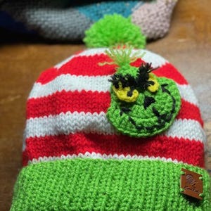 Peut inclure: Bonnet en tricot rayé rouge et blanc avec un pompon vert et un visage de Grinch en crochet vert sur le devant. Le bonnet a une bordure en tricot vert et une étiquette en cuir marron avec le texte "The Knitter" dessus.