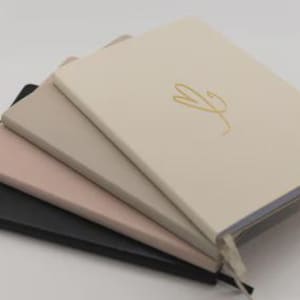 Puede incluir: Una pila de cuatro cuadernos en colores neutros: negro, rosa, gris y crema. El cuaderno crema superior tiene un diseño de escritura dorada. Cada cuaderno tiene una cinta de marcador.