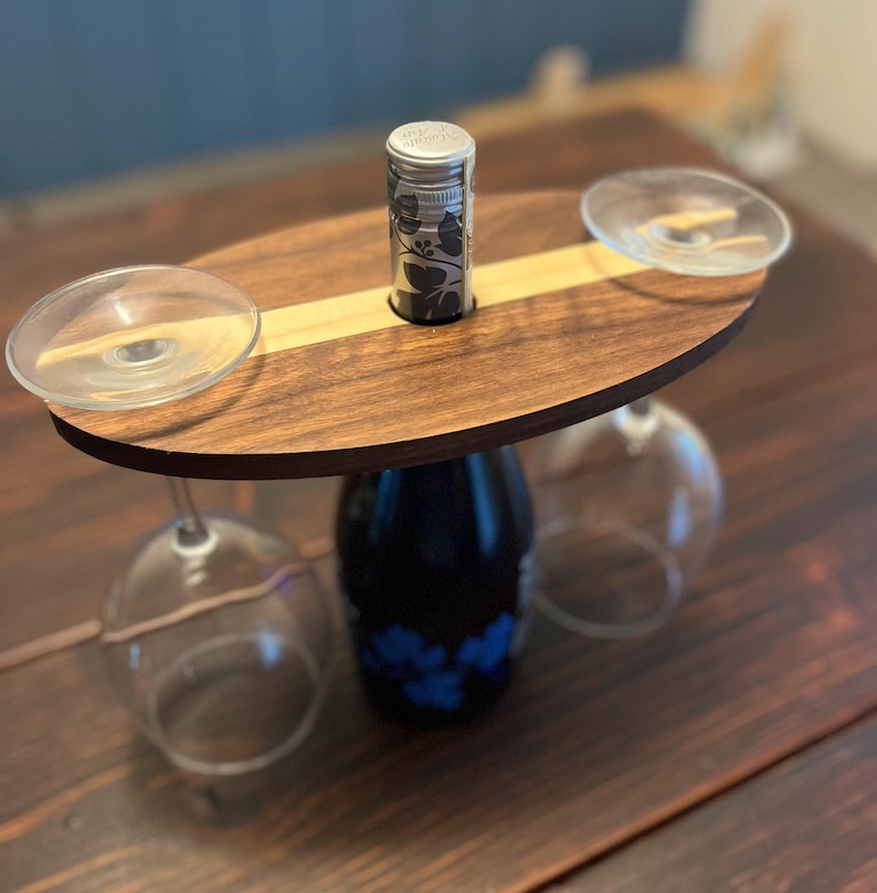 May include: A wooden wine caddy with a dark brown finish and a light wood stripe. It holds a wine bottle, two wine glasses, and a bottle stopper. The wine bottle is dark blue with a floral design.