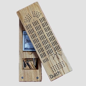 Op de afbeelding: Een houten cribbage-bord met een scharnierend deksel. Het deksel is open en toont het cribbage-bord met gaten voor pinnen. Het bord is gemaakt van licht hout met een donkere houtkleur. Het bord heeft het woord "Dutch" gegraveerd op het deksel.