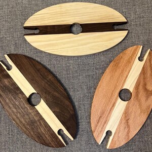 May include: Three wooden wine glass holders in oval shapes. Each has a central hole for the glass stem and a slot for the glass base. The holders are made of different wood types, including light, dark, and reddish-brown tones.