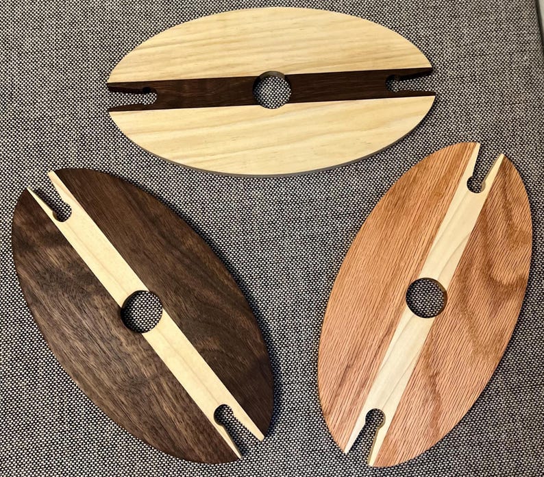 May include: Three wooden wine glass holders in oval shapes. Each has a central hole for the glass stem and a slot for the glass base. The holders are made of different wood types, including light, dark, and reddish-brown tones.