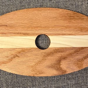 May include: Oval wooden wine glass holder with a light wood central section and a hole for the bottle. It features two cutouts for wine glasses. The wood grain is visible.