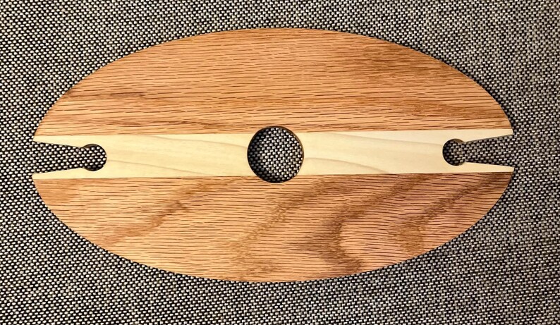 May include: Oval wooden wine glass holder with a light wood central section and a hole for the bottle. It features two cutouts for wine glasses. The wood grain is visible.