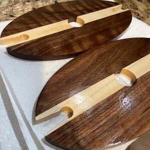 May include: Two oval wooden wine caddies. Each caddy is made of dark brown and light brown wood, with a circular hole and a notch for holding a wine glass. The caddies are on a white surface.