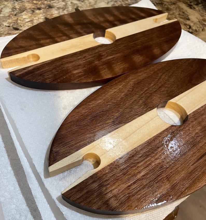 May include: Two oval wooden wine caddies. Each caddy is made of dark brown and light brown wood, with a circular hole and a notch for holding a wine glass. The caddies are on a white surface.