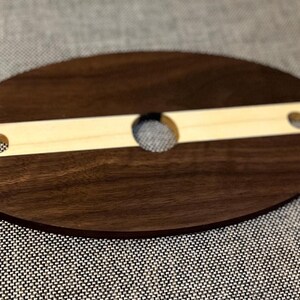 May include: A wooden wine glass holder. The oval base is dark brown, with a light-coloured wooden insert. The insert has a circular hole and two slots for holding wine glasses. The holder is designed to keep wine glasses stable.