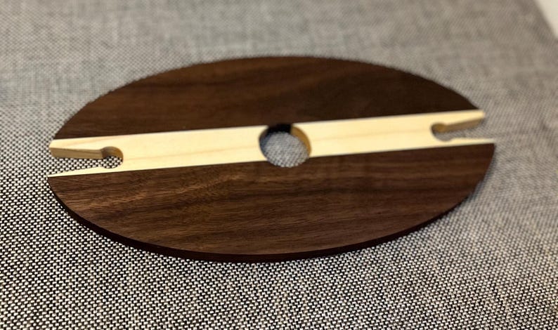 May include: A wooden wine glass holder. The oval base is dark brown, with a light-coloured wooden insert. The insert has a circular hole and two slots for holding wine glasses. The holder is designed to keep wine glasses stable.