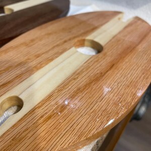 May include: Wooden serving board for wine and snacks, featuring a light wood central strip and two circular cutouts for wine glasses. The board is made of light brown wood with a natural grain.