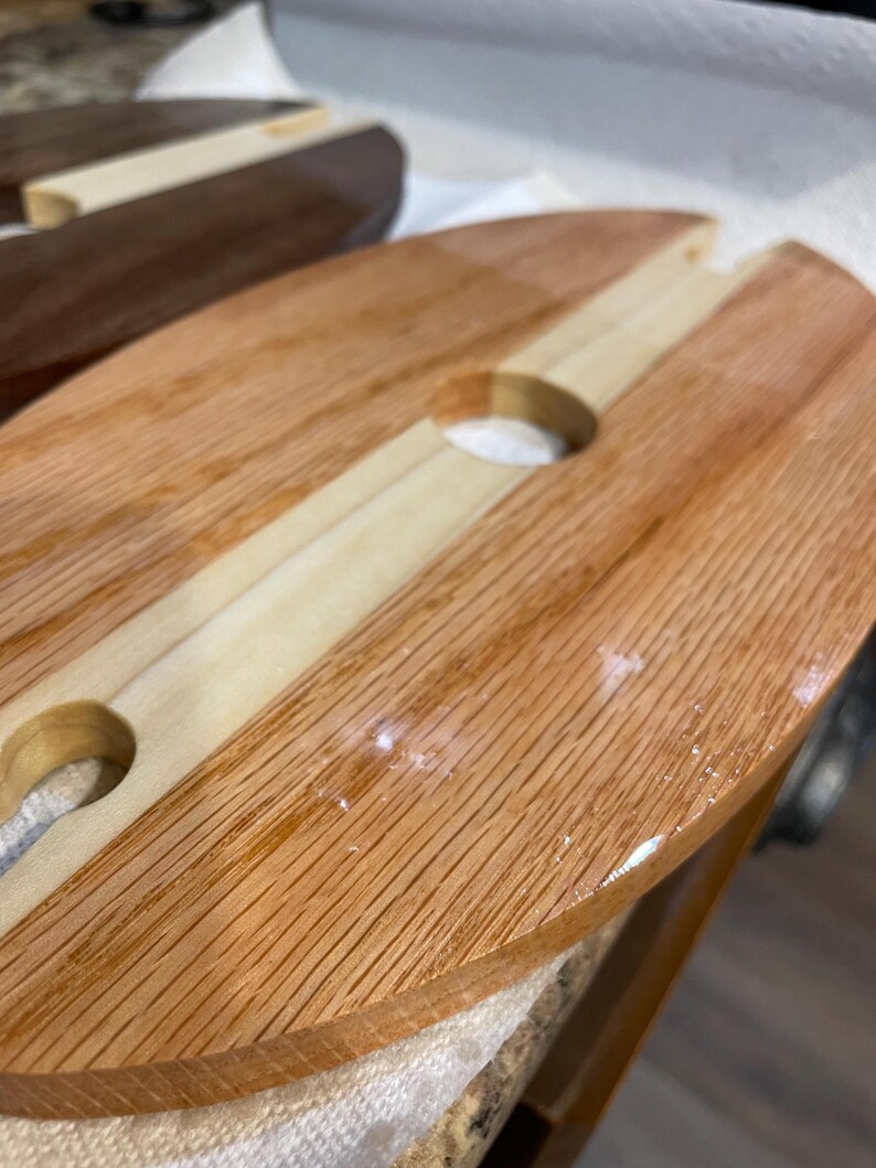May include: Wooden serving board for wine and snacks, featuring a light wood central strip and two circular cutouts for wine glasses. The board is made of light brown wood with a natural grain.
