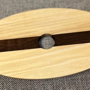 May include: Oval wooden wine glass holder with a light wood grain and a dark brown stripe. The holder has a circular opening in the centre and notches on either side to hold a wine glass. The holder is approximately 20 cm long.