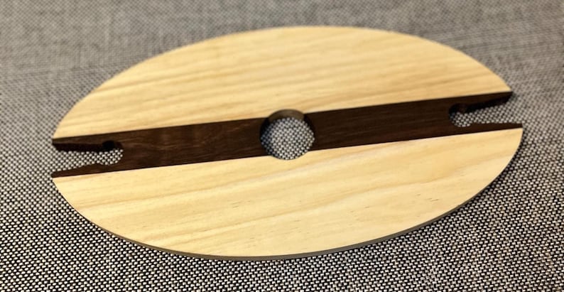 May include: Oval wooden wine glass holder with a light wood grain and a dark brown stripe. The holder has a circular opening in the centre and notches on either side to hold a wine glass. The holder is approximately 20 cm long.