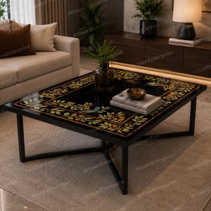 Op de afbeelding: Een zwarte vierkante salontafel met een decoratief bloemeninlegontwerp in goud en geel. De tafel heeft een zwart metalen frame en staat op een neutraal gekleurd tapijt. Een bank en andere meubels staan op de achtergrond.