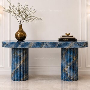 May include: A blue stone console table with a smooth, rounded rectangular top and two fluted, cylindrical bases. The surface features a marbled pattern with deep blue, white, and gold veining. A brass vase with branches and a stack of books sit on top.