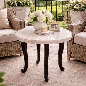 May include: A round marble-topped side table with dark brown legs. The table is set with a white vase of flowers, a candle, and a framed photo. Wicker chairs and potted plants flank the table, creating an outdoor living space.