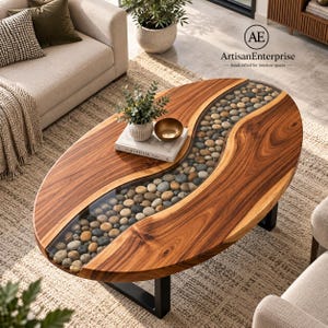 May include: An oval coffee table featuring a natural wood grain surface with a central, winding glass-encased channel filled with smooth, multi-colored river stones. The table is supported by black metal legs and holds a book and a small bowl.