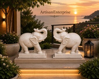 Puede incluir: Dos estatuas de elefantes de piedra blanca con las trompas levantadas, cada una sobre una base rectangular. El texto ArtisanEnterprise está centrado sobre las estatuas. Las esculturas están situadas en un patio de piedra con plantas.