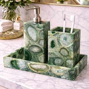 Puede incluir: Un juego de baño que incluye un dispensador de jabón, un soporte para cepillos de dientes y una bandeja. El juego está hecho de piedra de ágata verde con patrones blancos y verdes. El dispensador de jabón tiene una bomba plateada. Dos cepillos de dientes blancos están en el soporte.