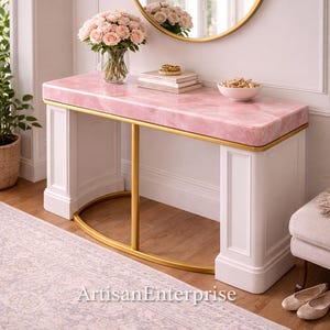 May include: A pink marble-topped console table with gold trim and white legs. A bouquet of pink roses sits in a glass vase on the table, along with books and a bowl. A gold-framed mirror hangs above the table. The text "Artisan Enterprise" is visible.