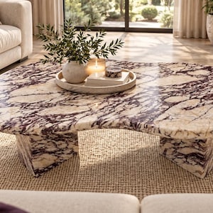 May include: A low-profile coffee table with an irregular, organic kidney-bean shape, crafted from polished cream-coloured marble featuring prominent, deep purple veining. The table is supported by two thick, matching marble slab legs.