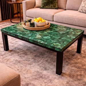 Puede incluir: Una mesa de centro rectangular con una vibrante tapa de malaquita verde y patas negras. La mesa está en un salón, con un sofá y una alfombra visibles. Una bandeja con limones y plantas está sobre la mesa.
