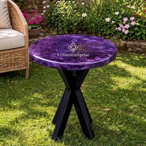 Puede incluir: Una mesa auxiliar redonda con una tapa morada y una base negra en forma de X. La tapa de la mesa tiene un aspecto veteado y el texto "ArtisanEnterprise" es visible. La mesa está colocada sobre un césped verde, con una silla de mimbre y un jardín al fondo.