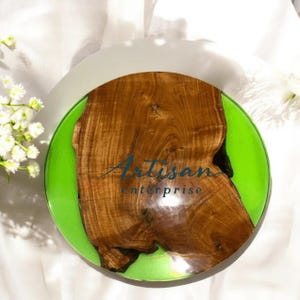 Może przedstawiać: Okrągła drewniana taca z zielonym brzegiem z żywicy epoksydowej. Taca ma naturalny wzór słojów drewna, a na drewnie nadrukowany jest tekst "Artisan enterprise".