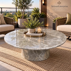 May include: A round coffee table with a polished grey marble top and a matching cylindrical marble base. On the table sits a gold circular tray holding a white book titled The Kinfolk Home, a small green potted plant, and a lit candle in a glass.