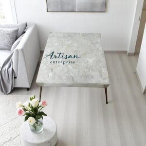 May include: A white marble coffee table with a grey and white marbled top. The table has a simple, modern design with thin metal legs. The table is sitting on a light wood floor. The text "Artisan enterprise" is printed on the table top.