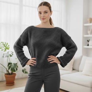 May include: A charcoal gray sweater with a boat neck and long sleeves. The sweater has a ribbed texture and is paired with matching gray pants. The model is standing in front of a window and a white couch.