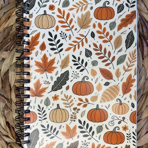 May include: A spiral-bound notebook with a fall-themed design. The cover features a white background with orange pumpkins, and various leaves in shades of orange, brown, and green. The notebook is resting on a woven, brown surface.