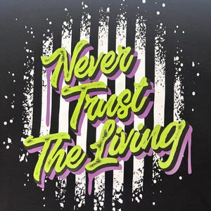 May include: A black t-shirt featuring a graphic print with the text Never Trust The Living in bright lime green with purple drop shadows. The background of the design consists of vertical white stripes with a distressed, paint-splattered effect.