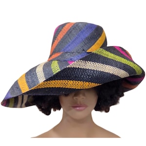 Puede incluir: Un sombrero de paja de ala ancha con un vibrante patrón de rayas multicolores. El sombrero muestra bandas alternas de negro, naranja, morado, verde lima y crema, tejidas en un diseño texturizado con una copa alta y un ala flexible e irregular.