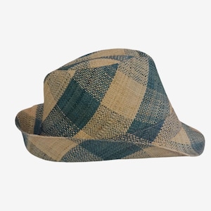 May include: A woven fedora hat featuring a prominent chequered pattern in alternating shades of natural beige and deep teal blue. The hat has a structured crown with a slight indentation and a curved brim that wraps around the base of the head.