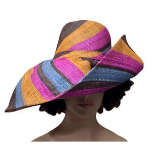 Puede incluir: Un sombrero de sol de ala ancha con textura tejida y rayas diagonales en tonos vibrantes de magenta, amarillo dorado, azul claro y marrón oscuro. El sombrero tiene una copa redondeada y un ala grande y asimétrica que se curva hacia arriba.
