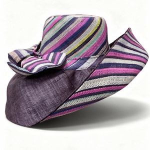 May include: A wide-brimmed hat featuring a woven texture with a multi-coloured striped pattern in shades of purple, pink, white, and yellow. The hat includes a large, decorative bow on the side and a solid dark purple section on the lower brim.
